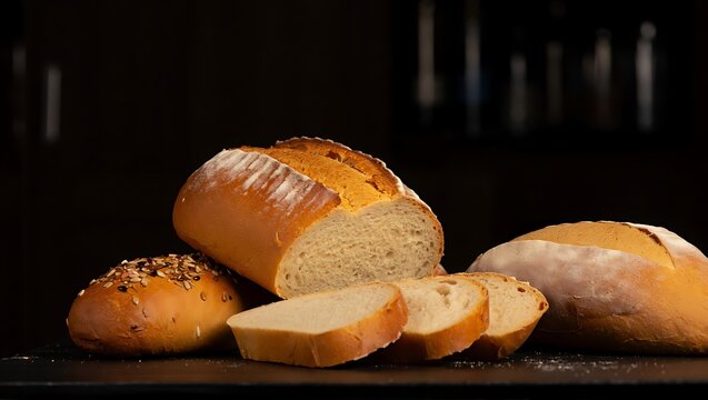 Freshly baked artisan bread, with one loaf sliced to reveal its golden crumb texture, positioned next to whole loaves and a seeded roll on a dark surface.