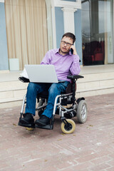 Young man in a wheelchair works at his laptop sitting against a beautiful sky.