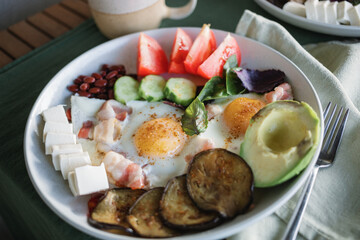 Close-up of a hearty homemade breakfast served on a large plate consisting of two fried eggs, bacon, feta cheese, avocado, tomatoes, eggplant, beans, basil leaves.