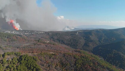 A high-angle view of a wildfire burning in a mountainous terrain, showcasing the smoke plume and surrounding landscape.