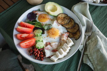 A hearty homemade breakfast served on a large plate consisting of fried eggs, bacon, feta cheese, avocado, tomatoes, eggplant, beans, basil leaves, on the garden table on the terrace.
