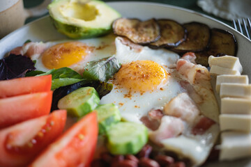 Close-up of a hearty homemade breakfast served on a large plate consisting of two fried eggs, bacon, feta cheese, avocado, tomatoes, eggplant, beans, basil leaves.