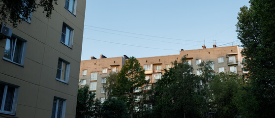 Wide-angle view of Soviet-era apartment buildings framed by lush green summer trees under clear...