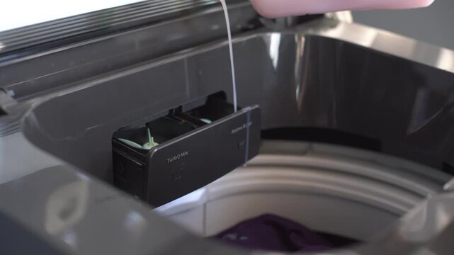 A woman's hand fills the dispenser of a top-loading washing machine with fabric softener.