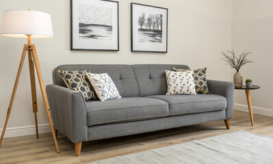 A modern living room setting featuring a light gray sofa with decorative pillows, positioned against a neutral wall. Above the sofa are two framed art pieces, and beside it stands a tall, wooden floor