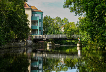 Fototapeta premium The Weiße Elster with buildings on the banks and the Peterssteg, a slightly arched steel arch bridge in Leipzig. The Elster cycle path runs over the Peterssteg. Reflection of the bridge in the water