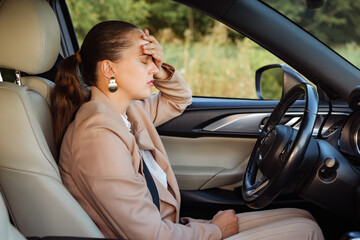 
A stressed and exhausted businesswoman sits in a car, with her hand on her forehead and eyes closed.
