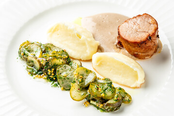 Plated pork tenderloin with creamy sauce, mashed potatoes, and pickled cucumber garnish. Traditional homemade meal served on a white decorative plate.
