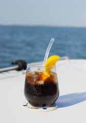Refreshing rum and cola cocktail with lemon slice, set on a white boat deck under a vivid blue sky. A perfect symbol of summer escape, relaxation, and seaside vibes. Crisp, cool, and carefree.