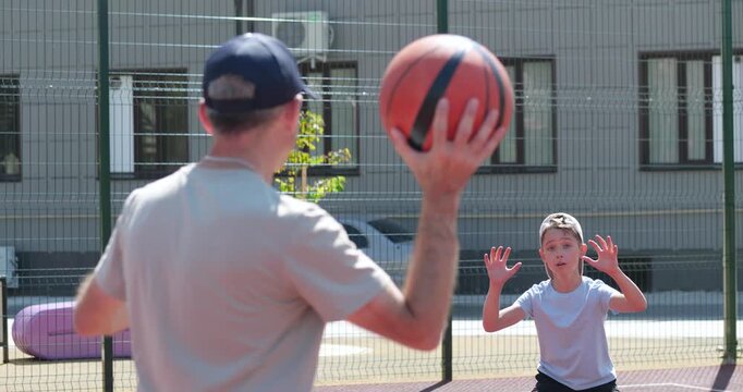 Father and son train basketball outdoors. Dad and son pass and catch ball with focus. Father and child enjoy active sports together, learning, bonding. Energetic father and boy practice. Joyful family