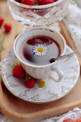 A beautiful cup of brewed tea sits on a decorative saucer, adorned with a daisy flower. Fresh strawberries complement the setting on a smooth wooden tray, creating a charming presentation