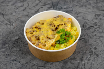 rice with vegetables and chop with mushrooms and cheese in a disposable plate on a gray background