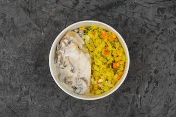 rice with vegetables and chicken fillet in a creamy sauce in a disposable plate on a gray concrete background