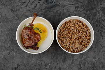 duck leg and buckwheat porridge in a disposable plate on a gray background with bread