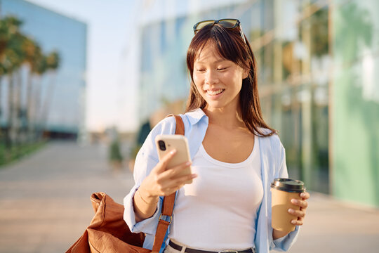 Smiling businesswoman walking through an urban street, chatting on her smartphone while enjoying a takeaway coffee during her morning commute - Powered by Adobe