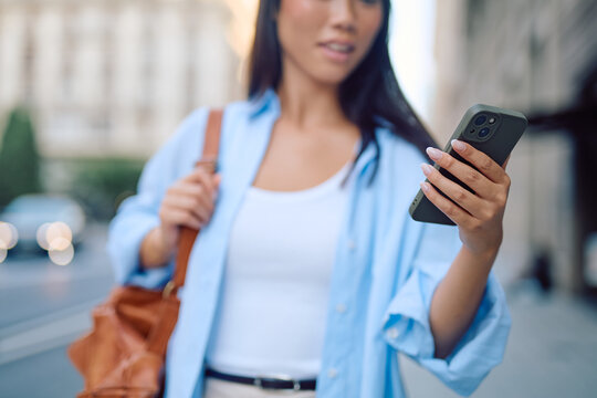 Asian businesswoman walking through an urban street, holding a smartphone and checking messages against a blurred city backdrop