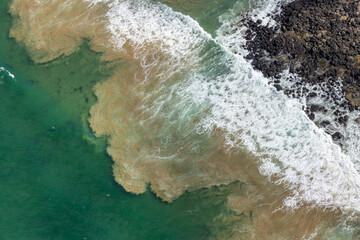 Aerial view of foamy waves crashing onto a dark, rocky shore where the sea transitions from deep green to a sandy brown near the coast, Breamlea, Victoria, Australia.