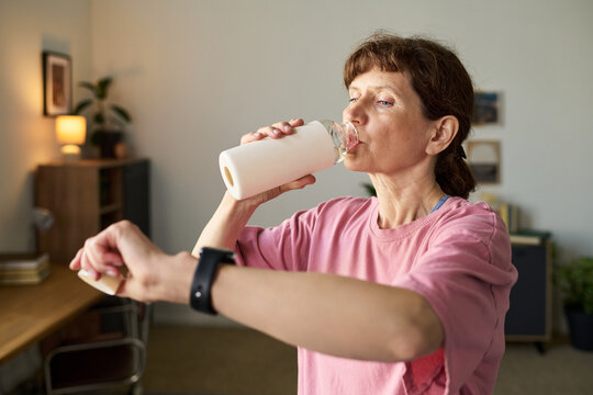 Senior Caucasian woman drinking from water bottle while checking smartwatch in home office setting, focusing on hydration and time management during daily routine