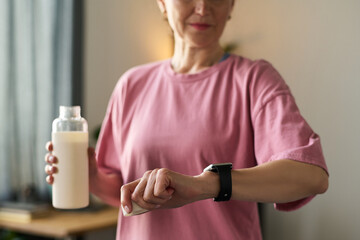 Senior Caucasian woman checking smartwatch while holding bottle, standing indoors with focus on hand gesture and wearable technology, face partially visible, casual setting