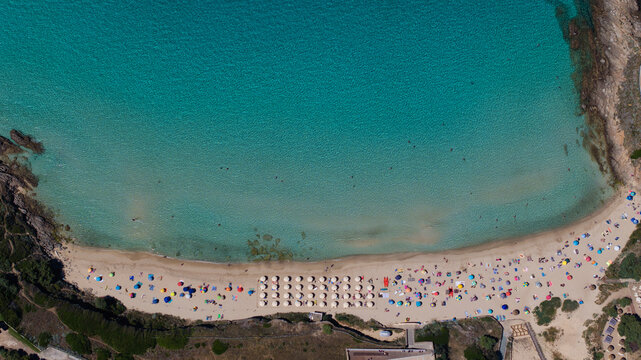 Aerial view of bright turquoise waters meeting the soft, sandy beach crowded with colorful umbrellas and sunbathers near Spiaggia La Pelosa, Stintino, Sardinia, Italy.