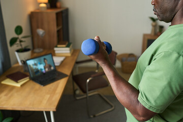 Senior Black man exercising with dumbbell while following online fitness class on laptop in home office, focusing on strength training and maintaining healthy lifestyle