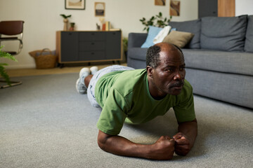 Senior Black man exercising in living room performing plank on carpeted floor, focusing on fitness and strength training with arms bent and body parallel to ground