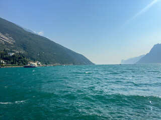 Fototapeta premium Scenic view of the turquoise waters of Lake Garda from a rocky promenade in Torbole, with mountains and buildings in the background. High quality photo