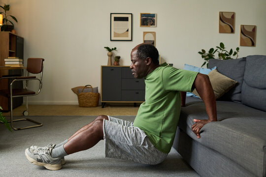 Senior Black man exercising at home performing triceps dips using sofa in living room, maintaining focused expression, demonstrating active lifestyle and strength training routine