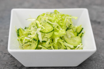 fresh cabbage and cucumber salad with greens in a white plate on a gray concrete background