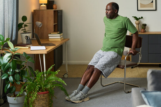 Senior Black man exercising by supporting body weight on arms while sitting on chair in home office setting, laptop and books on desk, indoor plants surrounding workspace