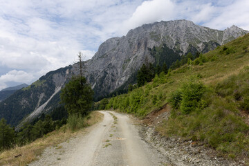 Gravel mountain road leading through green slopes with steep cliffs and dramatic rocky peaks in the background