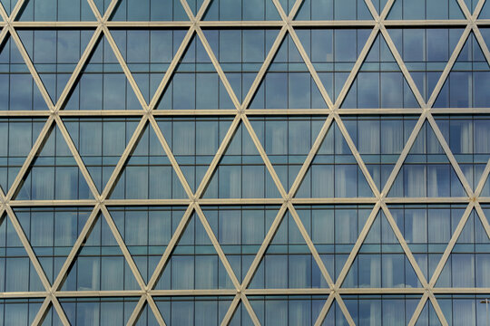 A building facade features a repeating pattern of triangular metal beams filled with reflective glass. The modern lattice design combines structural precision with visual rhythm.