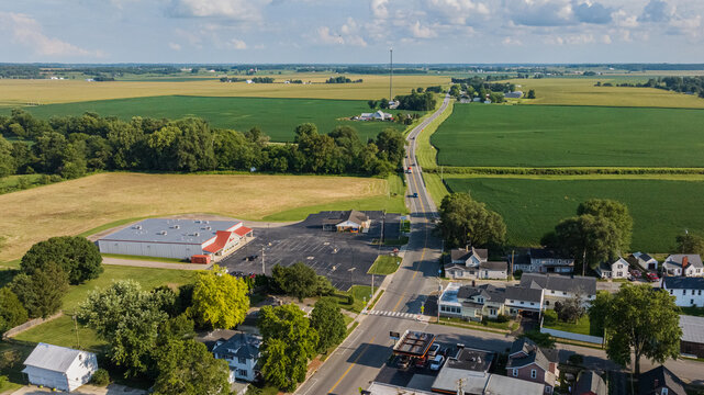 Aerial view of a road bisecting golden fields and lush green farmlands, complemented by buildings and houses nestled among trees, West Liberty, Ohio, United States.