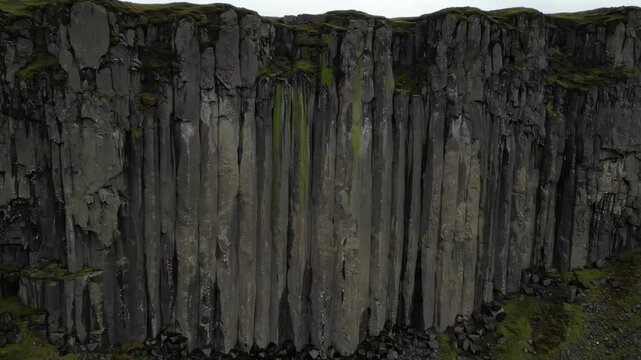 Aerial view of a dramatic basalt cliff formation under a cloudy sky, showcasing unique geological features, with lush greenery peeking through the rocky surfaces, ideal for nature documentaries