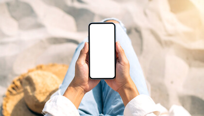 Overhead view of hands holding smartphone with blank screen, relaxing on sunny beach, summer vacation concept, mockup.