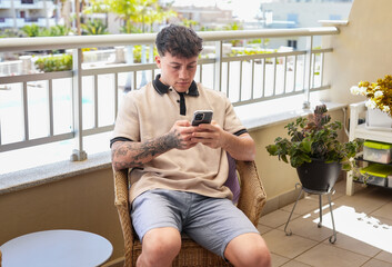Young caucasian smiling 20 years old boy focused on his smartphone sitting in outdoor terrace enjoying tech and social. Modern lifestyle, relaxation, technology use