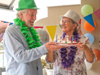 Joyful elderly couple celebrates a birthday in a festive and colorful setting. Wearing party hats they share a birthday cake marked with candles shaped “75,” surrounded by balloons and decorations