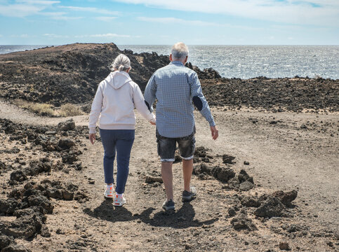 Senior couple walking together by the ocean, supporting each other, enjoying active aging and outdoor leisure on a scenic rocky coastline under a clear blue sky - Powered by Adobe