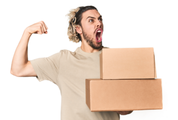 Young man with cardboard boxes in studio raising fist after a victory, winner concept.