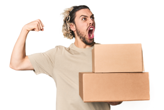 Young man with cardboard boxes in studio raising fist after a victory, winner concept.