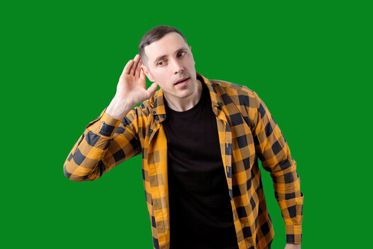 A young man in a yellow and black checked shirt stands against a green background. He raises his hand to his ear as if listening and looks away.