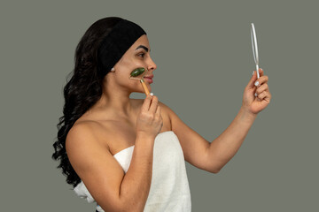 Obraz premium Woman using green jade roller and holding a mirror for facial massage