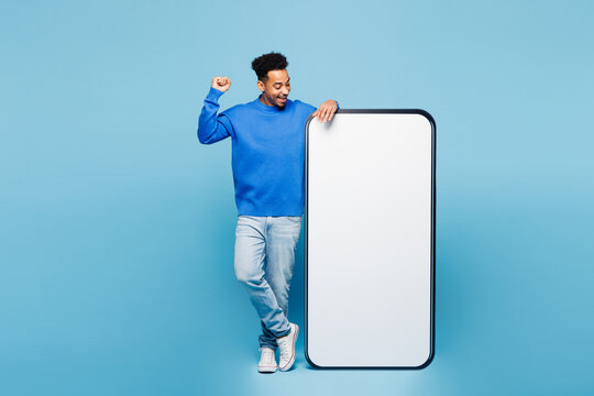 Full body young happy man of African American ethnicity wear sweatshirt casual clothes big huge blank screen mobile cell phone smartphone with area do winner gesture isolated on plain blue background.