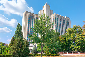 Obraz premium Stately government building in Chisinau stands tall behind hedges, symmetrical facade marked by vertical columns and a Moldovan flag fluttering above. Scene exudes civic formality and national pride