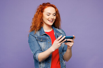 Young ginger woman wears red t-shirt denim shirt use play racing app on mobile cell phone hold gadget smartphone for pc video games isolated on plain pastel light purple background. Lifestyle concept.