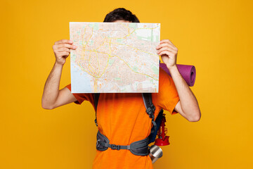 Young traveler man carry backpack with stuff mat reading paper map isolated on plain yellow background. Tourist leads active healthy lifestyle walk on spare time. Hiking trek rest travel trip concept.