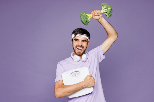 Young strong fitness trainer instructor man sportsman wear t-shirt training in home gym hold scales measure tape broccoli dumbbell isolated on plain purple background. Workout sport fit abs concept.