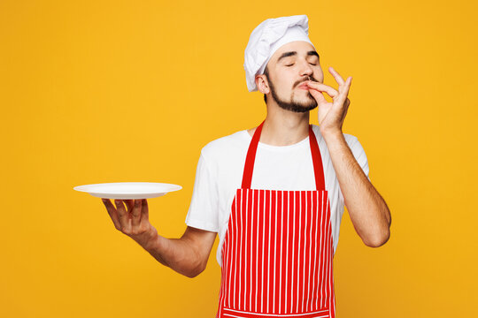 Young happy housekeeper chef cook baker man wear red apron toque hat hold in hand empty plate making okay taste delight sign, savoring isolated on plain yellow orange background. Cooking food concept. - Powered by Adobe