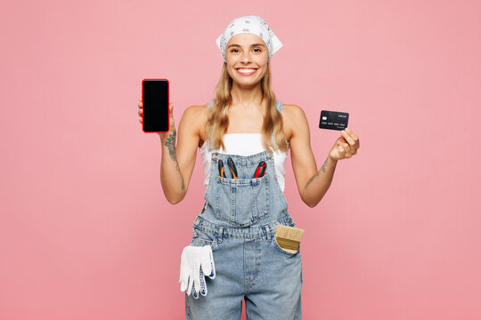 Young laborer handyman woman wear denim overhaul hold credit card use blank screen mobile cell phone isolated on plain pink background. Instruments for renovation apartment room. Repair home concept.