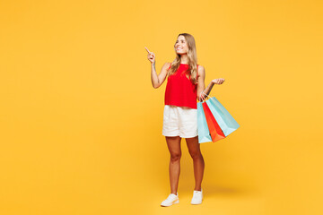 Full body young woman wear red casual clothes hold in hand shopping paper package bags point index finger aside on area isolated on plain orange yellow background. Black Friday sale buy day concept.